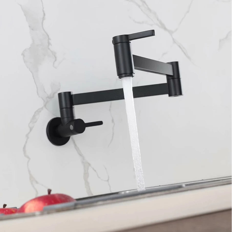 A matte black wall-mounted faucet with a single lever handle and a curved spout, shown with water running from the spout into a sink. The wall is white marble, and red apples are visible in the foreground.