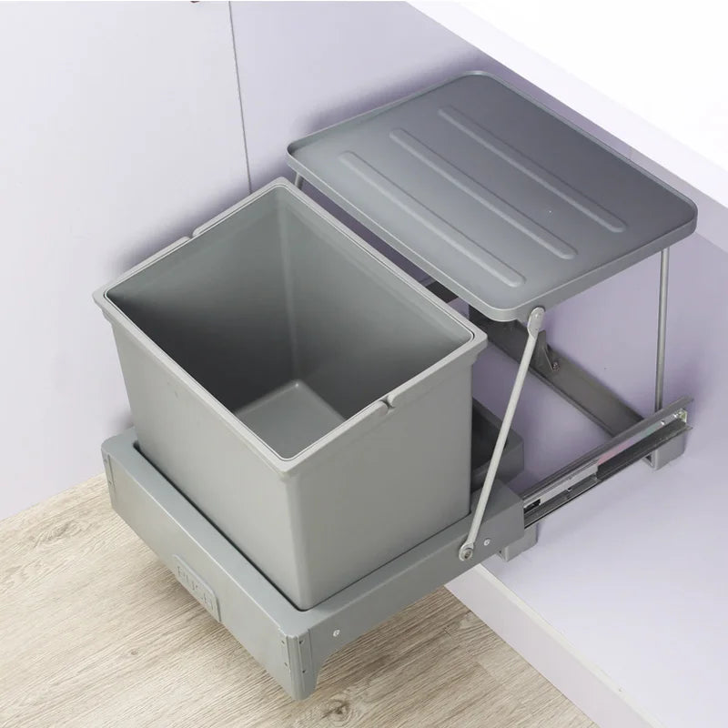 Gray pull-out trash bin with lid on a wooden floor against a white wall.
