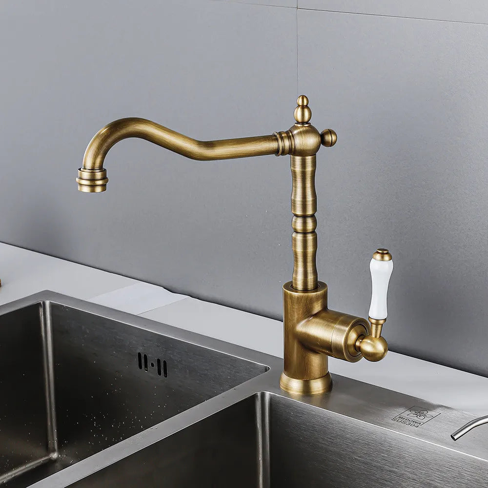 Brass kitchen faucet with white handle on a metallic sink against a gray wall.