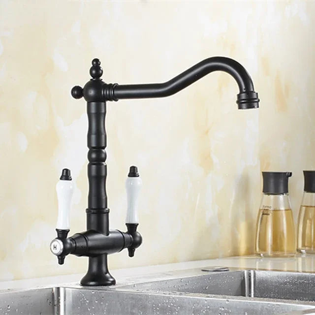 A black metal kitchen faucet with two white ceramic handles, mounted over a stainless steel sink against a beige tiled wall.