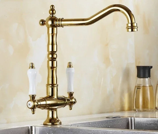 An antique brass kitchen faucet with two white ceramic handles, mounted over a stainless steel sink against a beige marble backsplash.