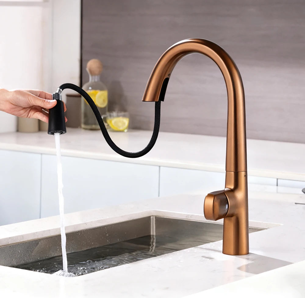 A modern kitchen faucet in a warm metallic copper finish with a black flexible pull-out sprayer. A hand is shown holding the sprayer to draw water from the sink, with a countertop and kitchen cabinets in the background.