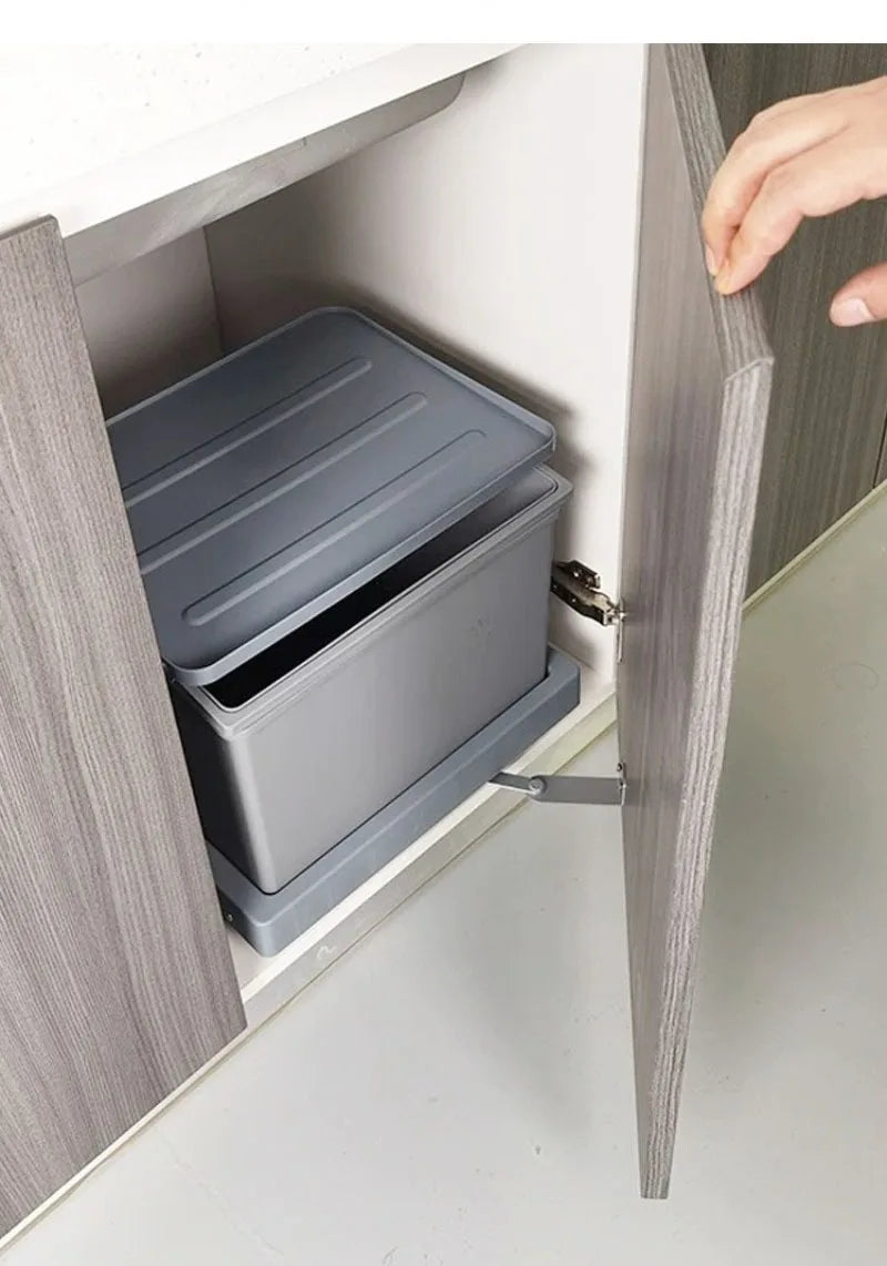 A gray plastic under-cabinet storage organizer box pulled out from a gray wood-look cabinet, with a hand opening the cabinet door.