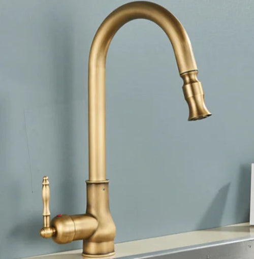 A single-handle kitchen faucet with a curved gooseneck spout in an antique brass or gold finish, mounted on a white sink against a light blue-gray wall.