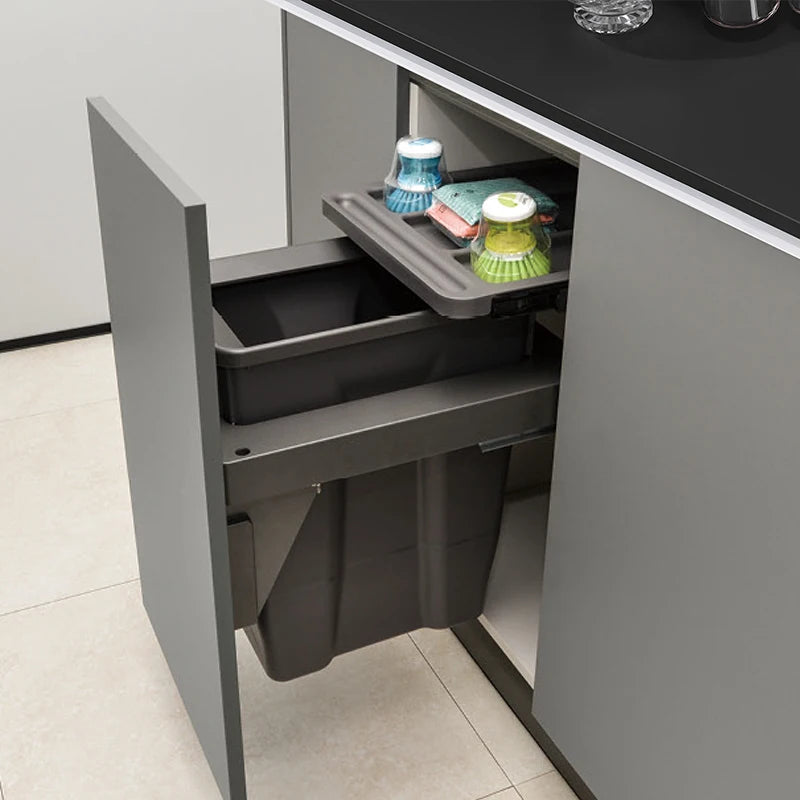 Grey kitchen cabinet with a pull-out trash bin and cleaning supplies on a tiled floor.