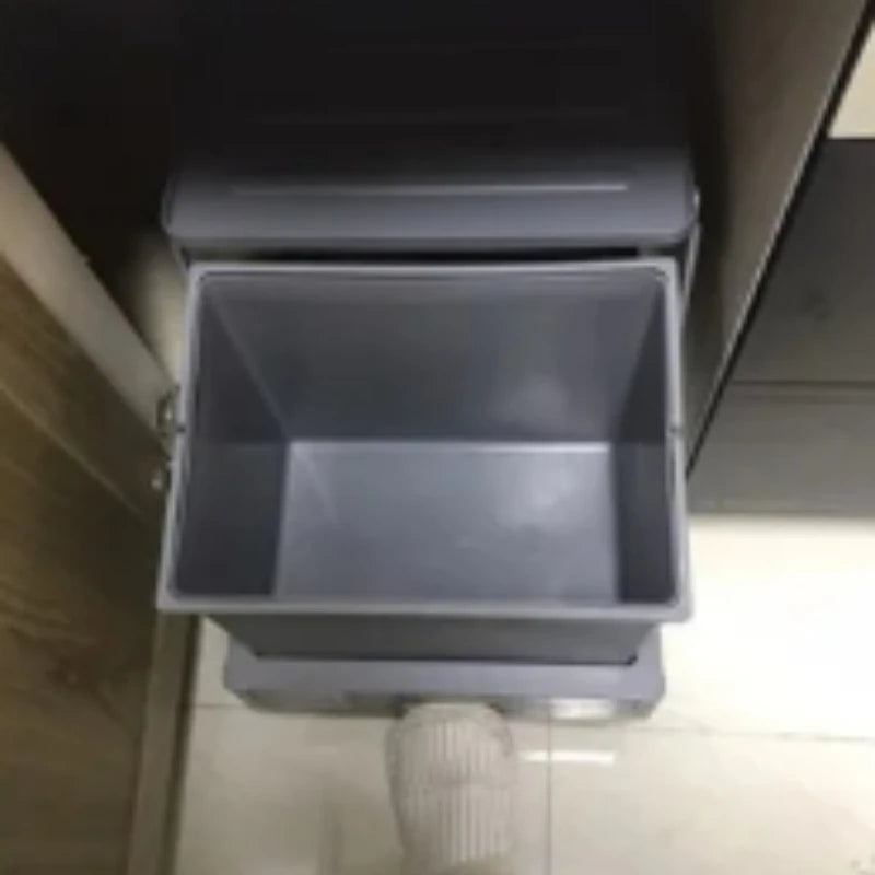 Gray plastic bin attached to a wall with a hose connected, on a tiled floor.
