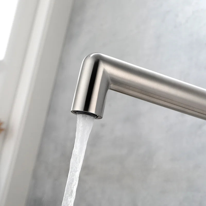 A close-up of a modern brushed nickel kitchen faucet with water flowing from the spout, set against a light gray textured wall.