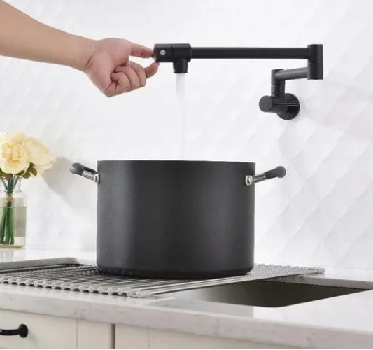 A black wall-mounted kitchen faucet with a long horizontal spout is shown with water running, while a large black pot sits on a metal rack over a sink. A human arm and hand are visible turning the faucet handle.