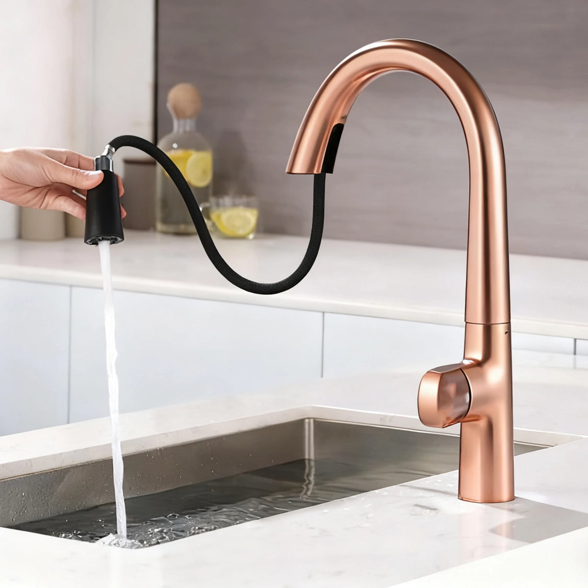 A copper-finished kitchen faucet with a curved high-arc spout and a detachable black flexible sprayer hose, shown with water running from the handheld sprayer into a sink. The faucet is installed in a bright modern kitchen.