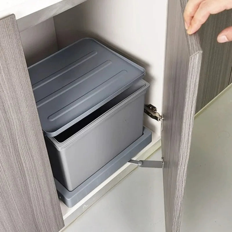 A gray plastic under-cabinet storage organizer box shown inside an open kitchen cabinet, with a hand opening the cabinet door.